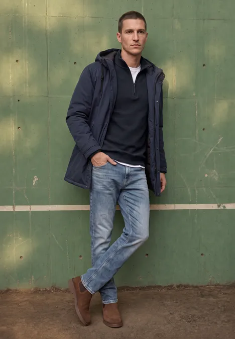 Ein Mann lehnt lässig in Jeans, braunen Boots und dunkelblauer Jacke an einer grünen Wand.