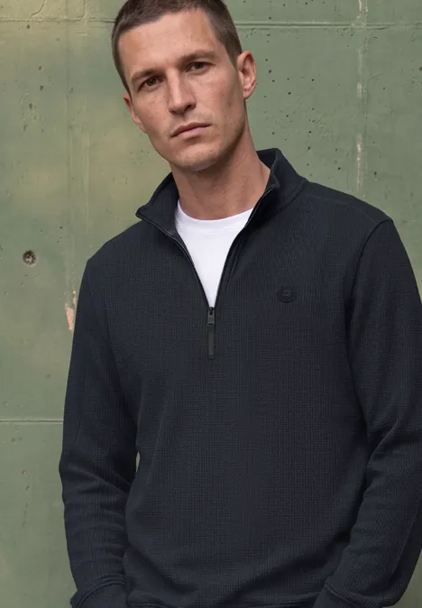 Ein Mann vor grünem Metallhintergrund trägt einen dunkelblauen Half-Zip-Pullover und ein weißes T-Shirt.