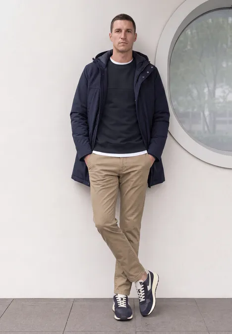 Ein Mann lehnt lässig an einer weißen Wand mit rundem Fenster und trägt Jacke, Pullover und Chinos.