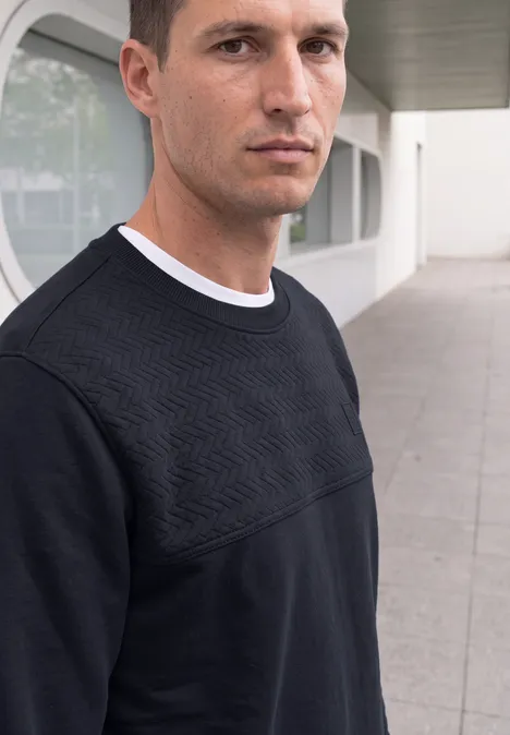 Ein Mann im schwarzen Pullover mit geometrischem Muster steht vor modernem Gebäude.