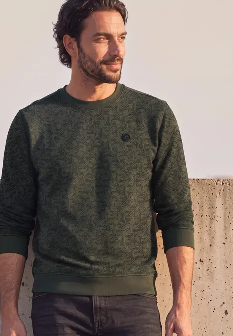 Sweat à col rond et imprimé urban olive green Mann mit dunkelgrünem Pullover, Bart und dunklen Haaren, steht seitlich vor einer Mauer im Freien.