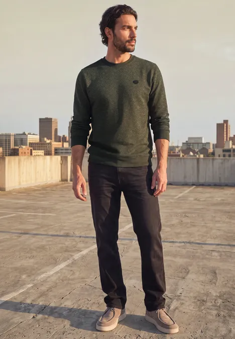 Sweat à col rond et imprimé urban olive green Mann in grünem Pullover und dunklen Hosen steht auf Parkdeckdach mit Stadtblick, schaut seitlich.