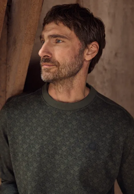 Sweat à col rond et imprimé urban olive green Ein bärtiger Mann in einem dunkelgrünen Pullover mit Muster blickt seitlich vor eine rustikale Wand.