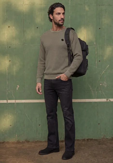 Ein Mann in grünem Pullover, dunklen Jeans und Rucksack steht vor einer grünen Wand.