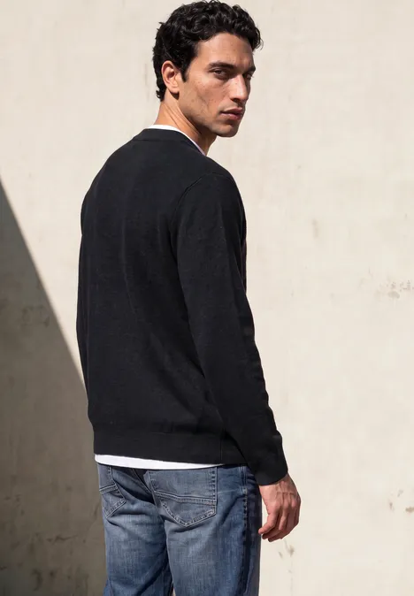 Ein junger Mann steht vor einer hellen Wand und trägt einen schwarzen Pullover und blaue Jeans.