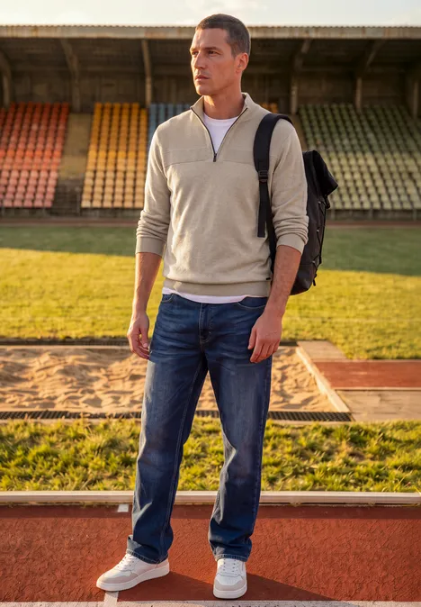 Junger Mann in Jeans und Pullover mit Rucksack steht auf der Stadion-Laufbahn neben dem Sandkasten.
