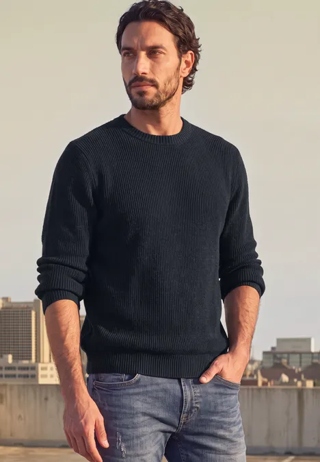 Ein Mann mit dunklem Pullover und Jeans auf einem Dach mit Stadtblick.