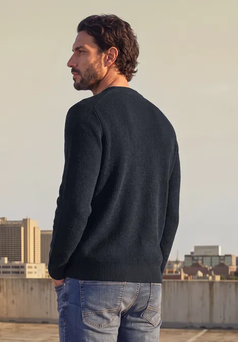 Ein Mann mit dunkelblauem Pullover und Jeans steht auf einem Dach und blickt auf die Stadt.