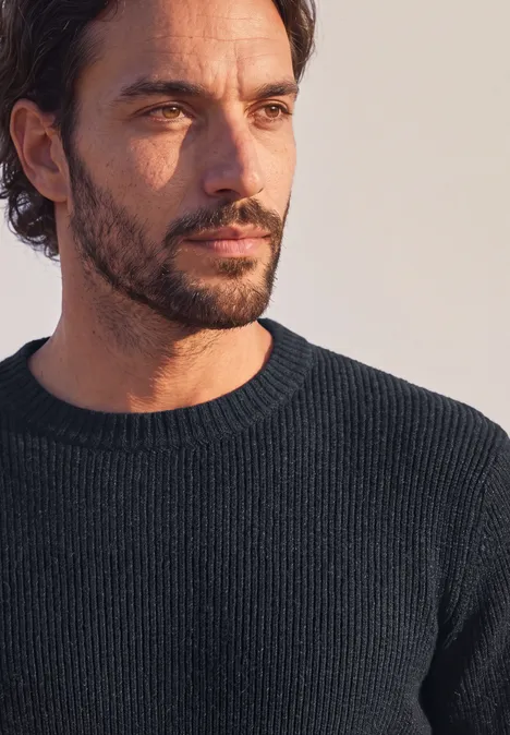 Nahaufnahme eines nachdenklich wirkenden Mannes mit dunklem Pullover, Bart und leicht gewelltem Haar.
