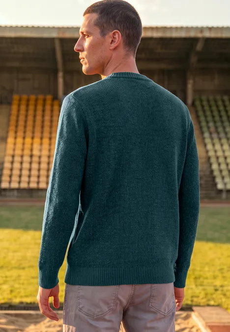 Pull en chenille à col rond dark fjord blue Mann im grünen Pullover steht mit dem Rücken zur Kamera vor leeren Tribünen im Freien.