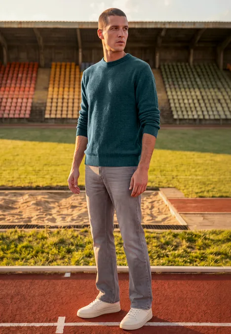 Pull en chenille à col rond dark fjord blue Ein Mann im Pullover und Jeans steht auf einer Laufbahn vor leeren Stadionrängen und schaut zur Seite.