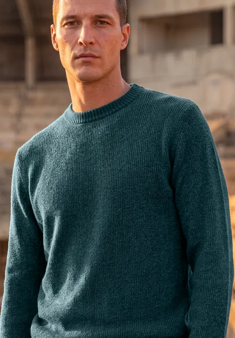 Pull en chenille à col rond dark fjord blue Ein Mann in einem dunkelgrünen Strickpullover steht vor einer unscharfen, urbanen Kulisse.