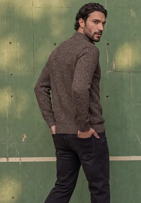 Pull à col roulé et texturé shadow brown Ein Mann in braunem Pullover und schwarzer Hose steht seitlich vor einem grünen Tennisplatz und blickt zurück.