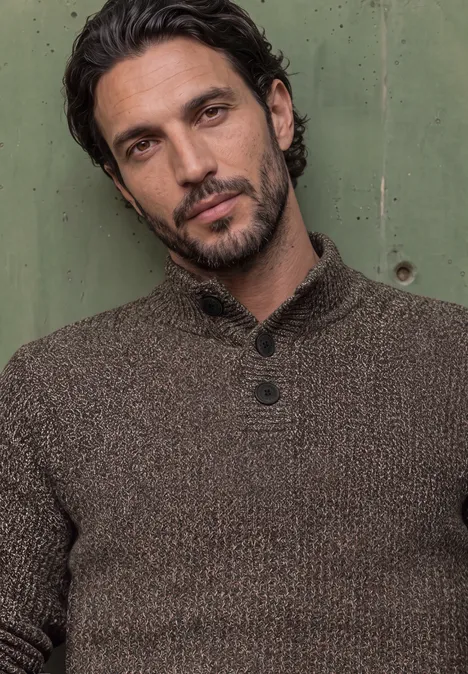 Pull à col roulé et texturé shadow brown Ein Porträt eines Mannes mit dunklen Haaren und Bart vor grünem Hintergrund, brauner Pullover.