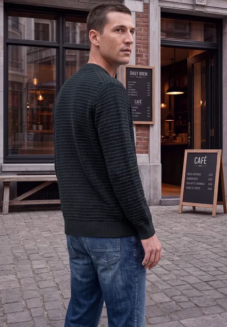 Chenille-trui met wafelstructuur dark ink blue Ein Mann in Jeans und Pullover blickt über die Schulter auf einen gepflasterten Weg vor einem Café.