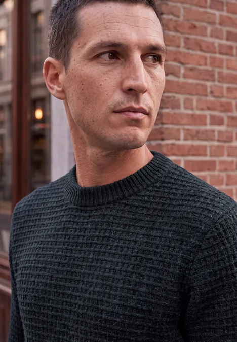 Chenille-trui met wafelstructuur dark ink blue Ein Mann mit kurzem dunklem Haar und schwarzem Strickpullover schaut seitlich vor einer Backsteinwand.