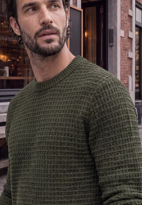 Chenille-trui met wafelstructuur urban olive green Mann mit olivgrünem, grob gestricktem Pullover vor städtischem Gebäude