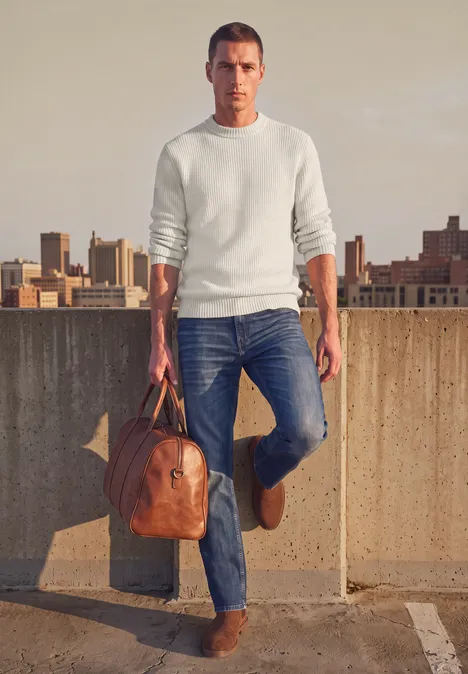 Ein Mann in Jeans, weißem Shirt und Ledertasche steht auf einem Parkdeck vor einer Stadtkulisse.