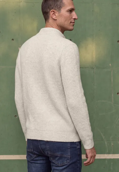Ein Mann in hellem Pullover und Jeans steht mit dem Rücken zur Kamera vor einer grünen Wand.