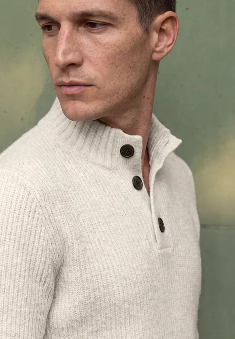 Ein Mann in einem hellen, gerippten Pullover schaut nach links vor einem grünen Hintergrund.