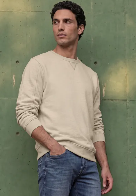 Sweat-shirt en tissu éponge avec détails en tricot côtelé canvas beige Un homme en pull clair et jean, appuyé contre un mur vert, regardant sur le côté.
