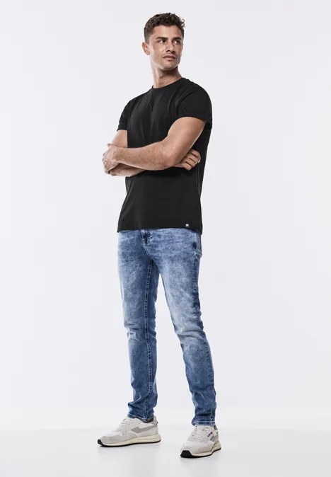 Homme en t-shirt noir, jean bleu et baskets blanches, bras croisés, debout.