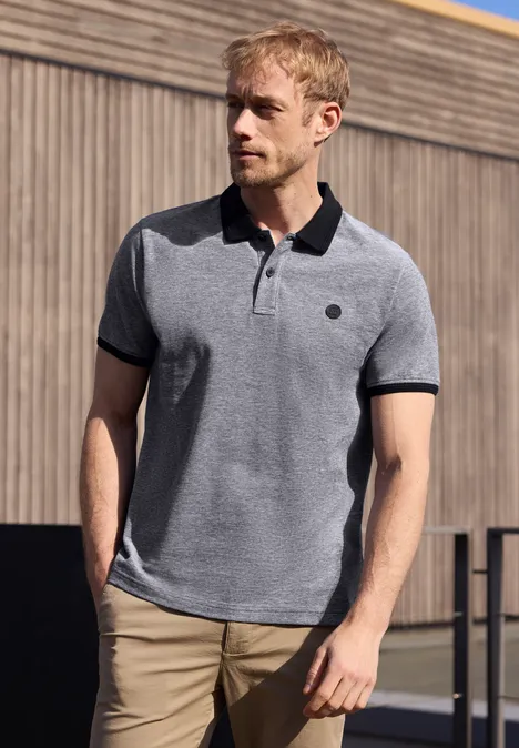 Een man staat voor een houten wand, draagt een grijs polohemd met zwarte kraag en beige broek.
