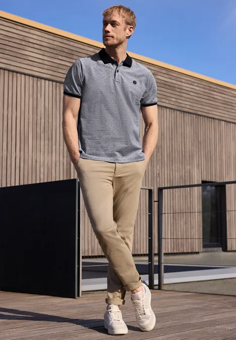 Een man in een grijs polo-shirt en beige chino’s staat voor een modern gebouw en kijkt in de verte.