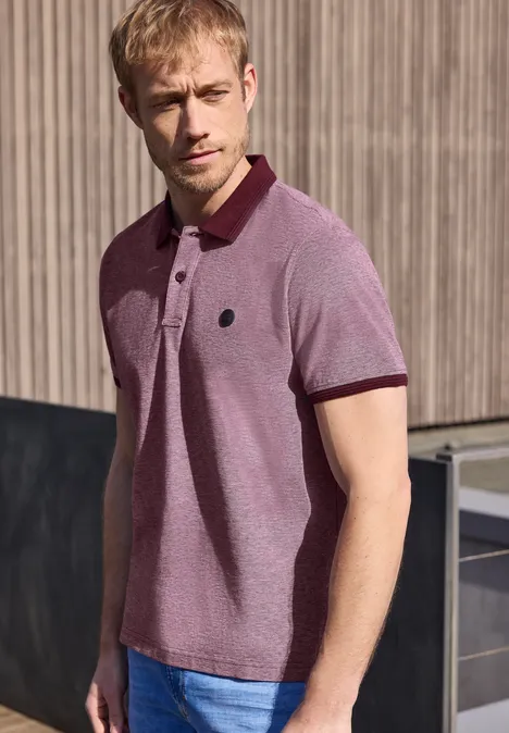 Een man in een roze poloshirt staat tegen een houten muur met blik opzij.