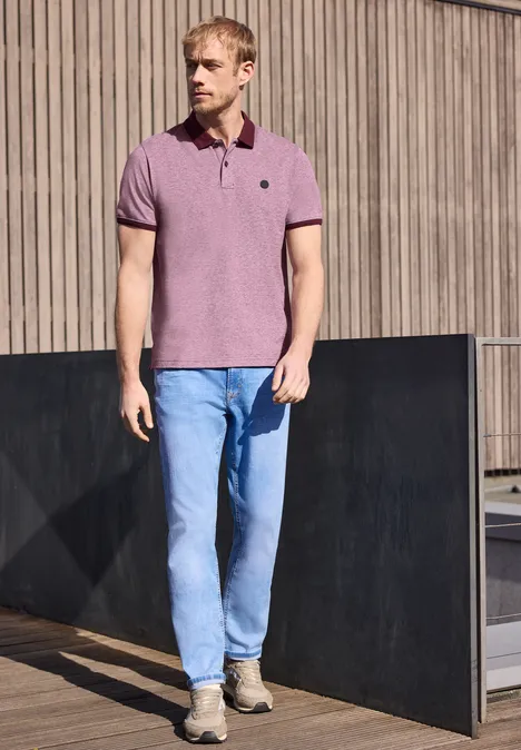 Een man in een roze poloshirt en blauwe spijkerbroek staat voor een houten gevel.