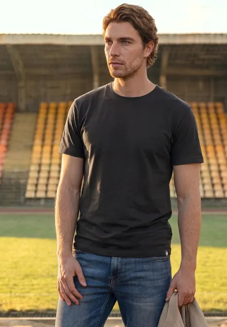 Un homme en t-shirt noir et jean se tient sur le terrain devant des gradins vides et regarde sur le côté.