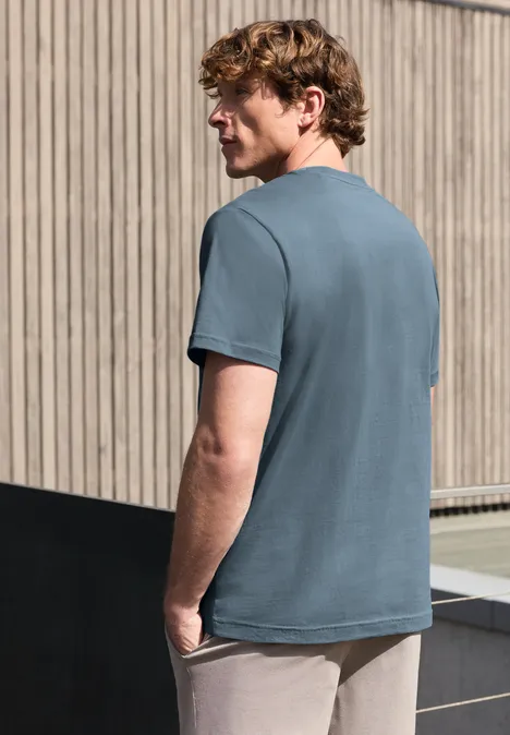 Een man in een blauwgrijs T-shirt met de rug naar de kijker, voor een houten gevel.