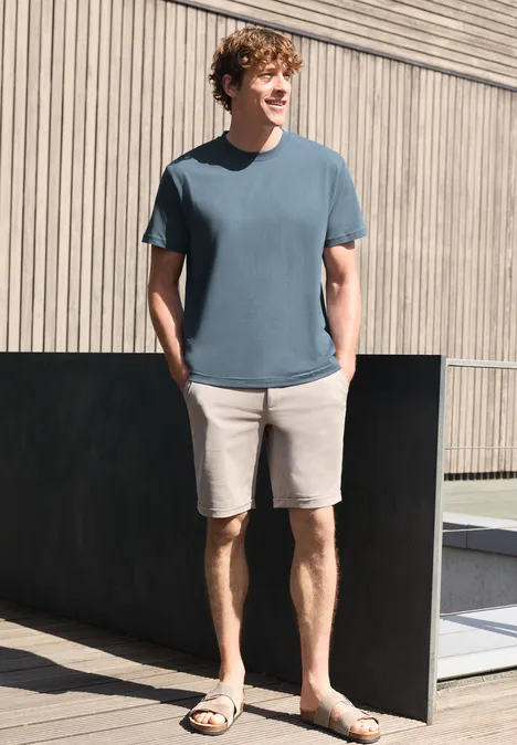 Een man draagt een blauw t-shirt, beige short en sandalen, staat ontspannen voor een houten gevel.