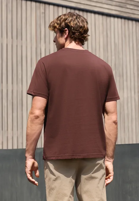Een man van achteren tegen een houten gevel, bruine t-shirt en beige broek