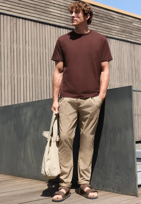Een man staat buiten voor een houten gevel, draagt een bruin t-shirt, lichte broek en sandalen, en houdt een tas vast.