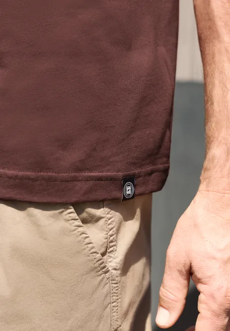 Close-up van een persoon met een bruin t-shirt en beige korte broek, rechter arm en hand zichtbaar.