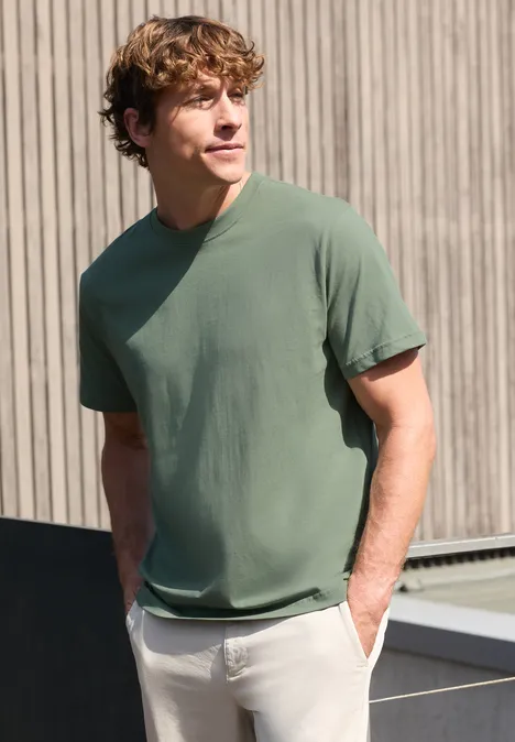 Een man met krullend haar draagt een groen t-shirt en staat ontspannen voor een houten gevel.