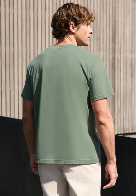 Een man met zijn rug naar de camera, gekleed in een groen shirt en lichte broek voor een houten gevel.