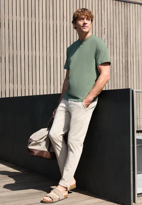 Een man leunt tegen een muur, draagt een groen T-shirt, lichte broek, sandalen en heeft een reistas.