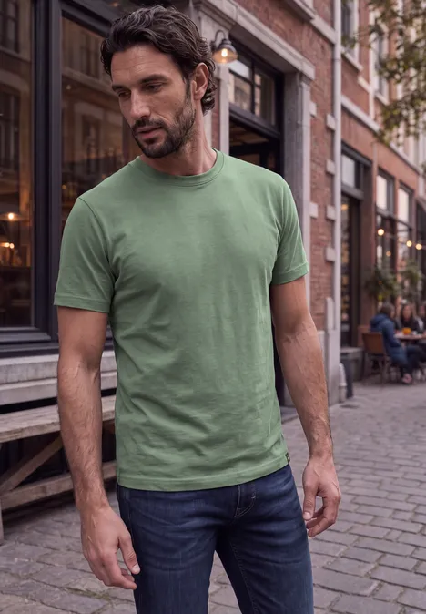 Ein bärtiger Mann in grünem T-Shirt und Jeans schlendert an einem Stadthaus mit Backsteinfassade.