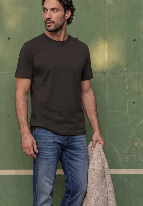 Basic Shirt van puur katoen shadow brown Man voor een groene achtergrond met donker T-shirt, spijkerbroek en houdt een beige jas vast.