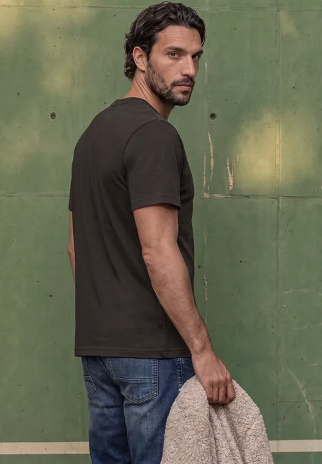 Basic Shirt van puur katoen shadow brown Een man met baard in zwart T-shirt en jeans kijkt over zijn schouder voor een groene muur en houdt een doek vast.
