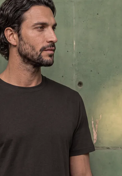 Basic Shirt van puur katoen shadow brown Een man met donkere baard en t-shirt kijkt naar rechts voor een groene muur.