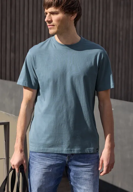 Een man staat voor een houten wand, draagt een blauw T-shirt, jeans en houdt een tas vast.