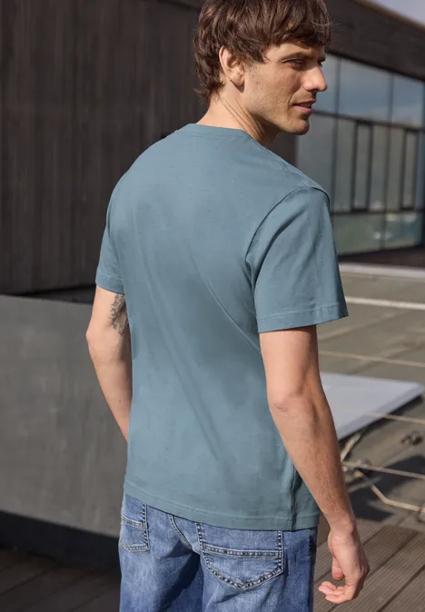 Een man op een terras, van achteren gefotografeerd, met een blauwgroen T-shirt en jeans, kijkt over zijn schouder.