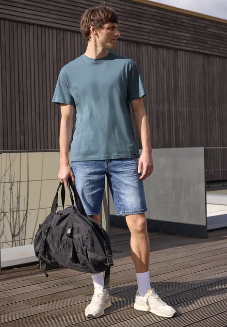 Een man staat op een houten terras, draagt een blauw T-shirt, jeansshorts, witte sneakers en houdt een tas vast.