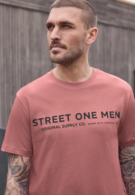 Ein Mann steht vor einem Holzzaun und trägt ein rosafarbenes T-Shirt mit dem Aufdruck „STREET ONE MEN“.