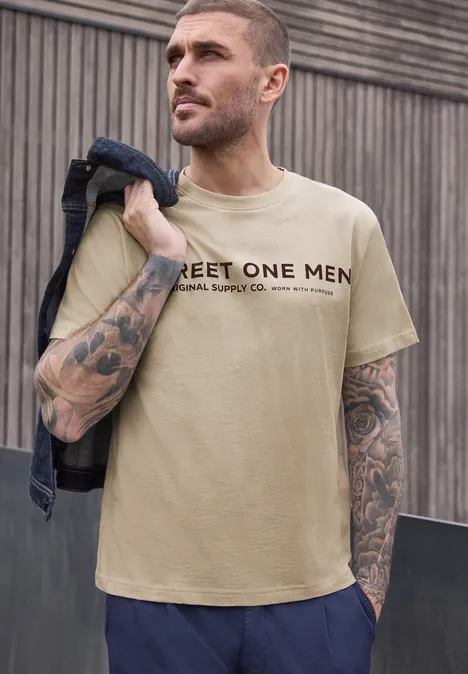 Mann mit beige T-Shirt 'STREET ONE MEN' und Denimjacke über der Schulter vor Holzwand