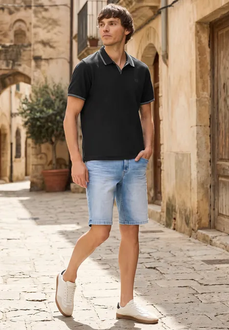 Ein Mann in schwarzem Poloshirt und Jeansshorts spaziert durch eine gepflasterte Gasse einer alten Stadt.