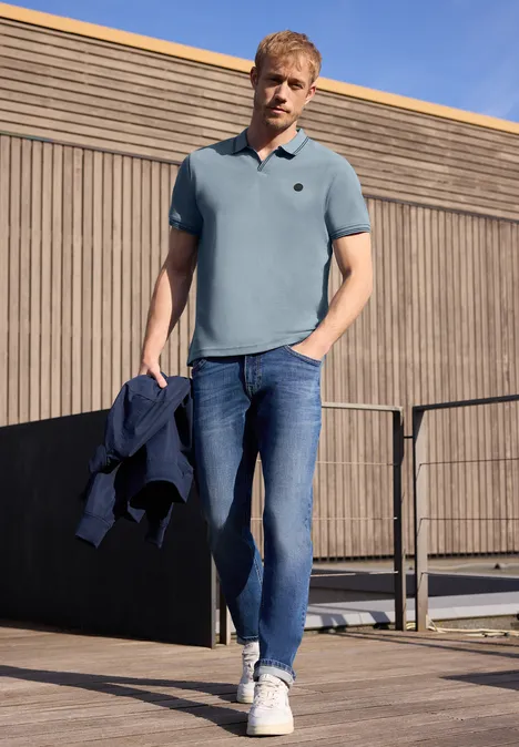 Ein Mann in blauem Poloshirt, Jeans und Sneakern steht auf einer Holzterrasse und hält eine Jacke in der Hand.
